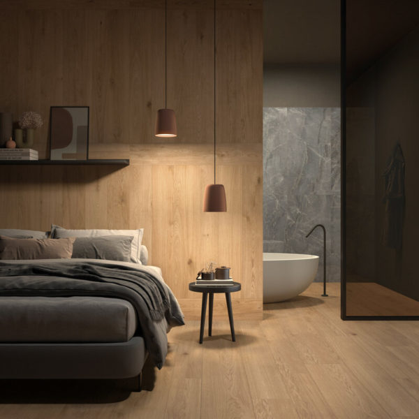Example Furnishing Of A Bedroom With OAK Cinnamon Parquet Flooring, A Double Bed Without Wooden Frame Cropped On The Left In The Picture, Next To It A Stool About As High As The Bed With Decorative Objects On It, In The Background The Passage To A Gray Marbled Bathroom With A White Bathtub Visible In The Cropped View